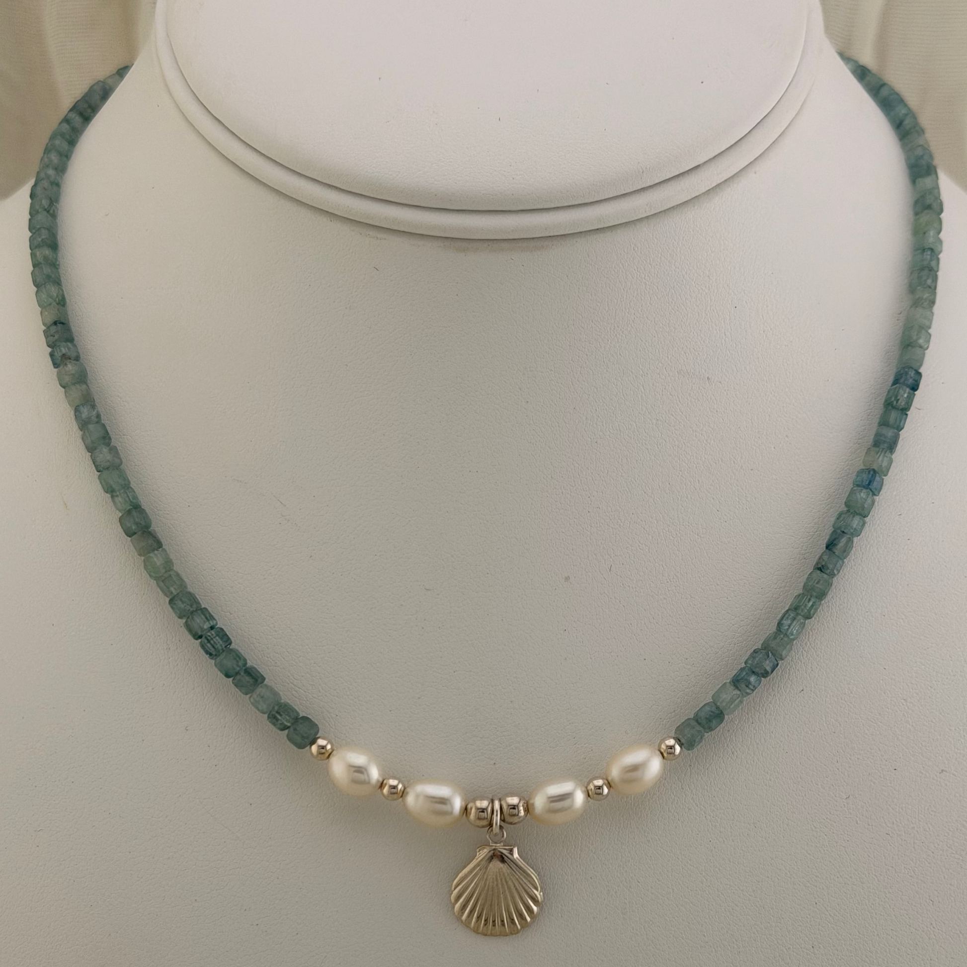 A gemstone beaded necklace displayed on a white mannequin bust, featuring a strand of teal-blue faceted beads, four white oblong pearls, small sterling silver spacer beads, and a sterling silver seashell pendant.