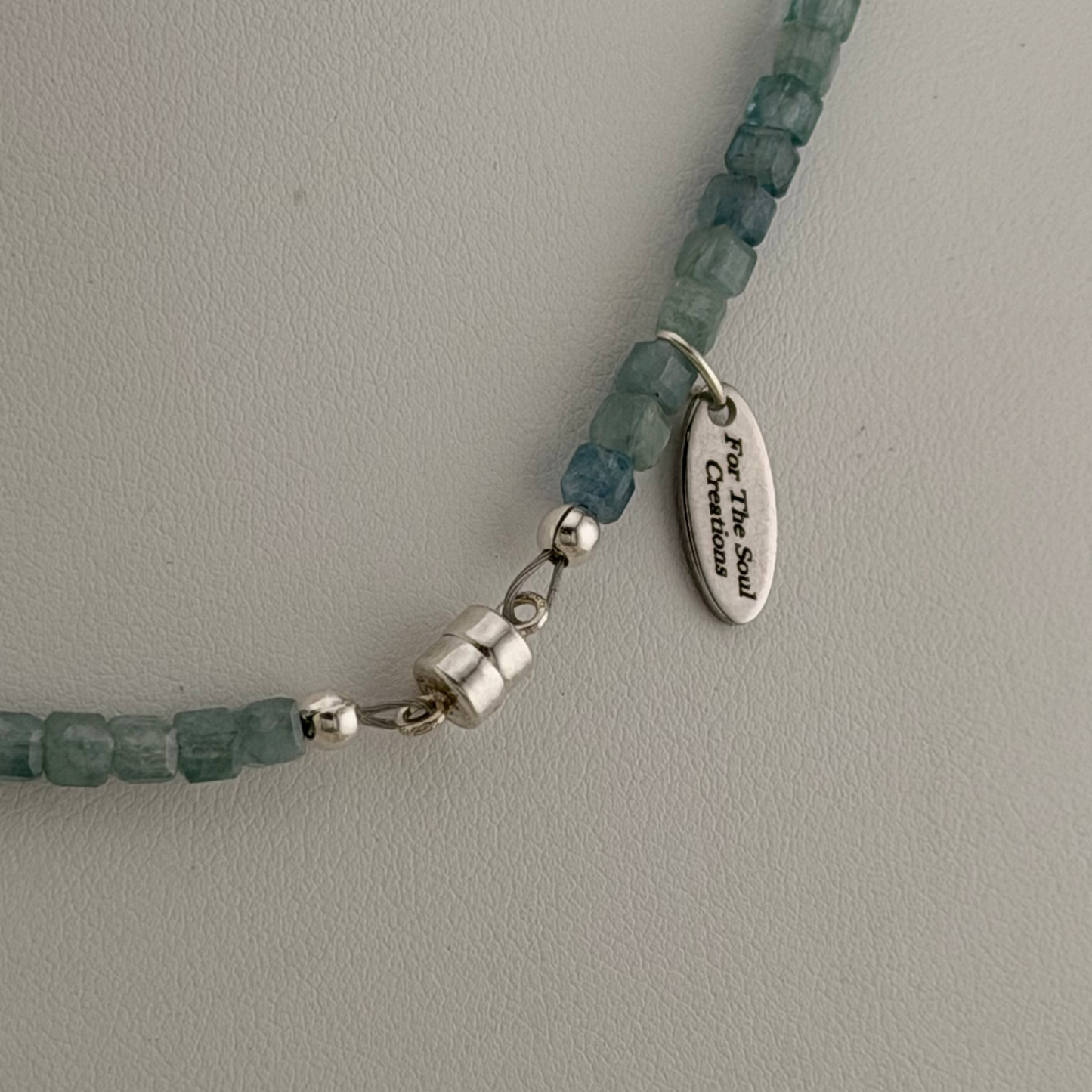 A close-up of a gemstone beaded necklace with faceted teal-blue beads and silver-toned components, including a small oval charm engraved with 'For The Soul Creations' and a silver magnetic clasp, laid on a light gray textured surface.