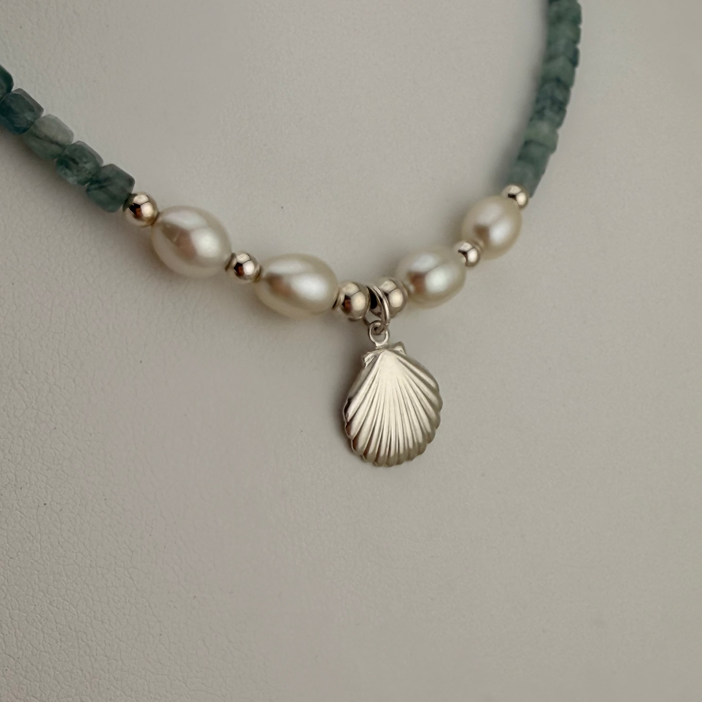 A closeup of gemstone beaded necklace featuring white freshwater pearls, small metallic silver spacer beads, and dark green faceted beads, with a silver-toned scallop shell pendant hanging from the center.
