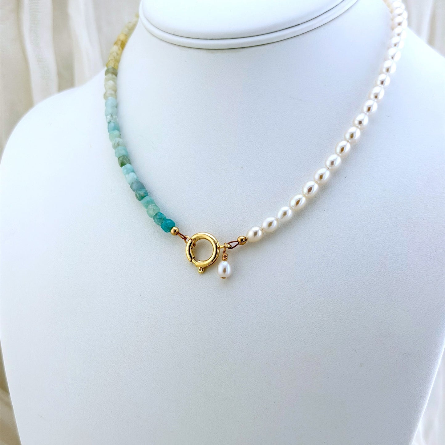 A delicate necklace displayed on a white jewelry bust, featuring a strand of white pearls and a side of aquamarine beads in light blue and pale yellow, finished with a gold clasp and a small pearl drop.