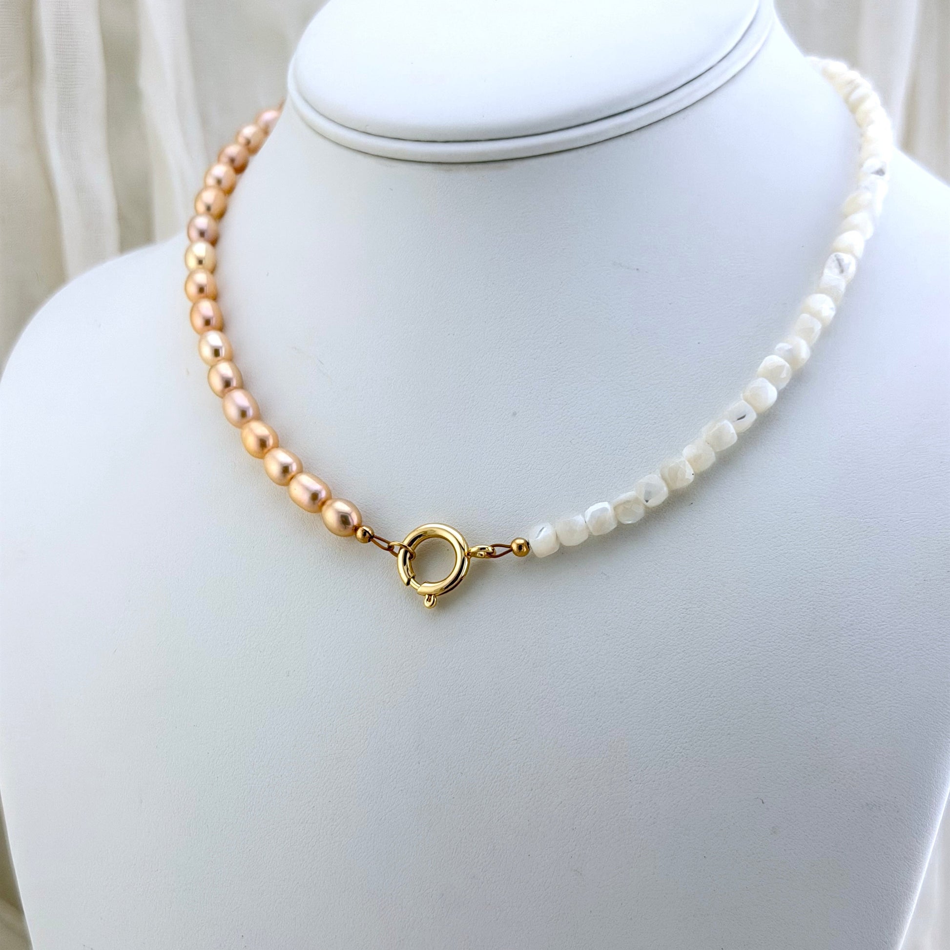 A delicate necklace displayed on a white jewelry bust, featuring champagne pink pearls on one side, mother-of-pearl beads on the other, and a gold clasp.