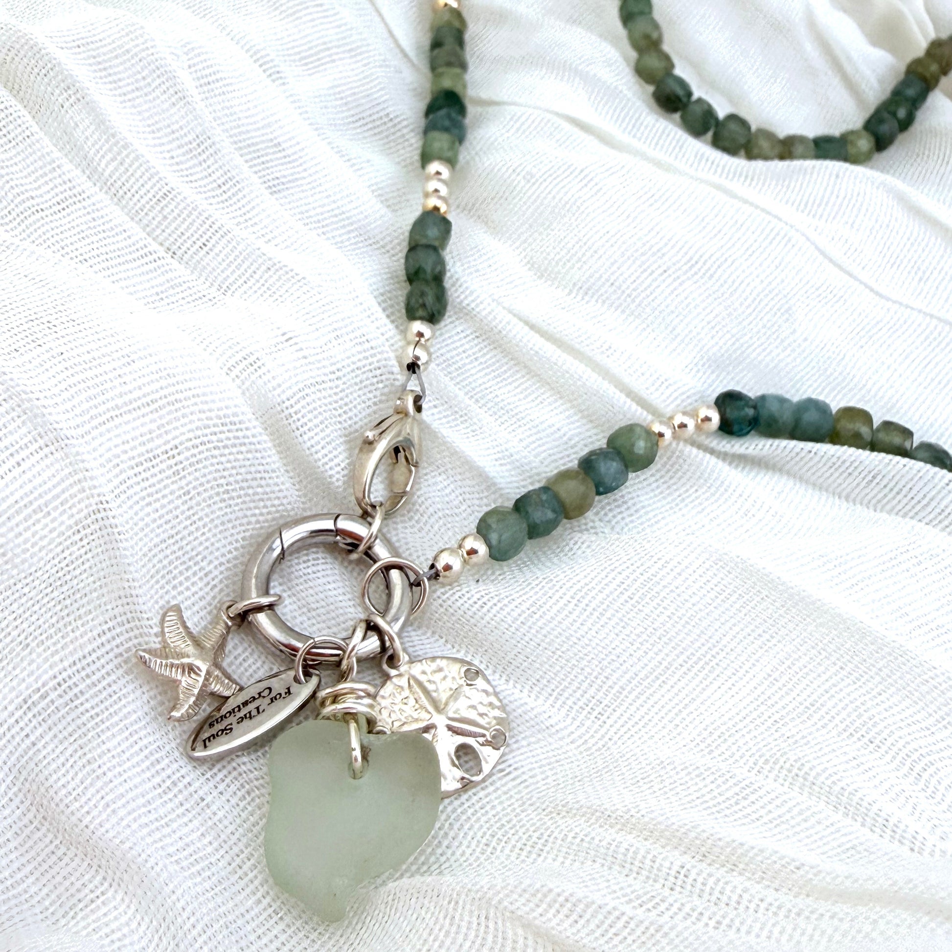 A close-up of a beaded necklace with green and silver beads, featuring a silver-toned ring clasp, silver beads, and  charms including a piece of seaglass, starfish, and sand dollar laid on white textured fabric.