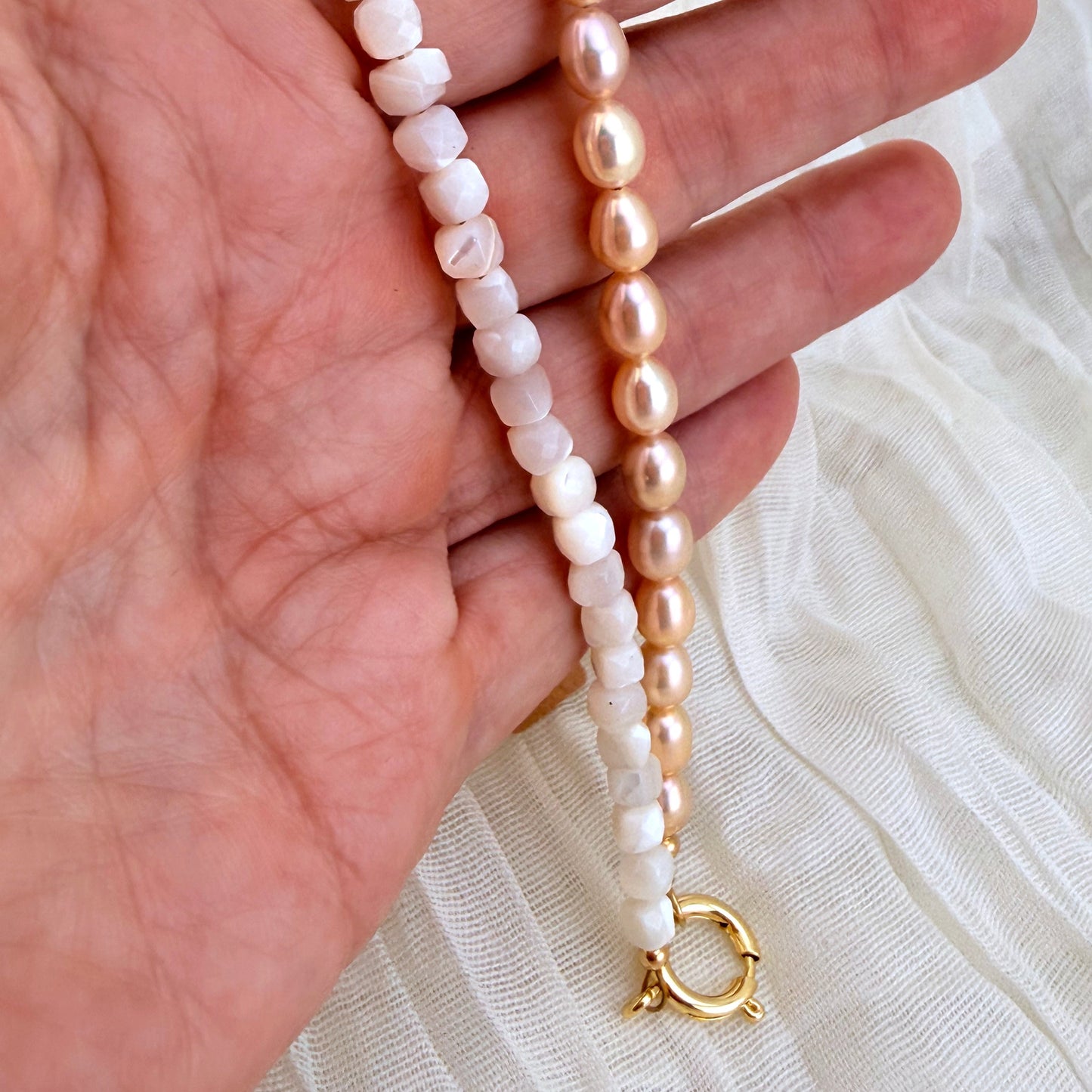 Two beaded strands draped over a hand: one strand features  white beads with gold settings, and the other features smooth, oval light pink pearls with gold settings. Both strands terminate in a gold clasp.