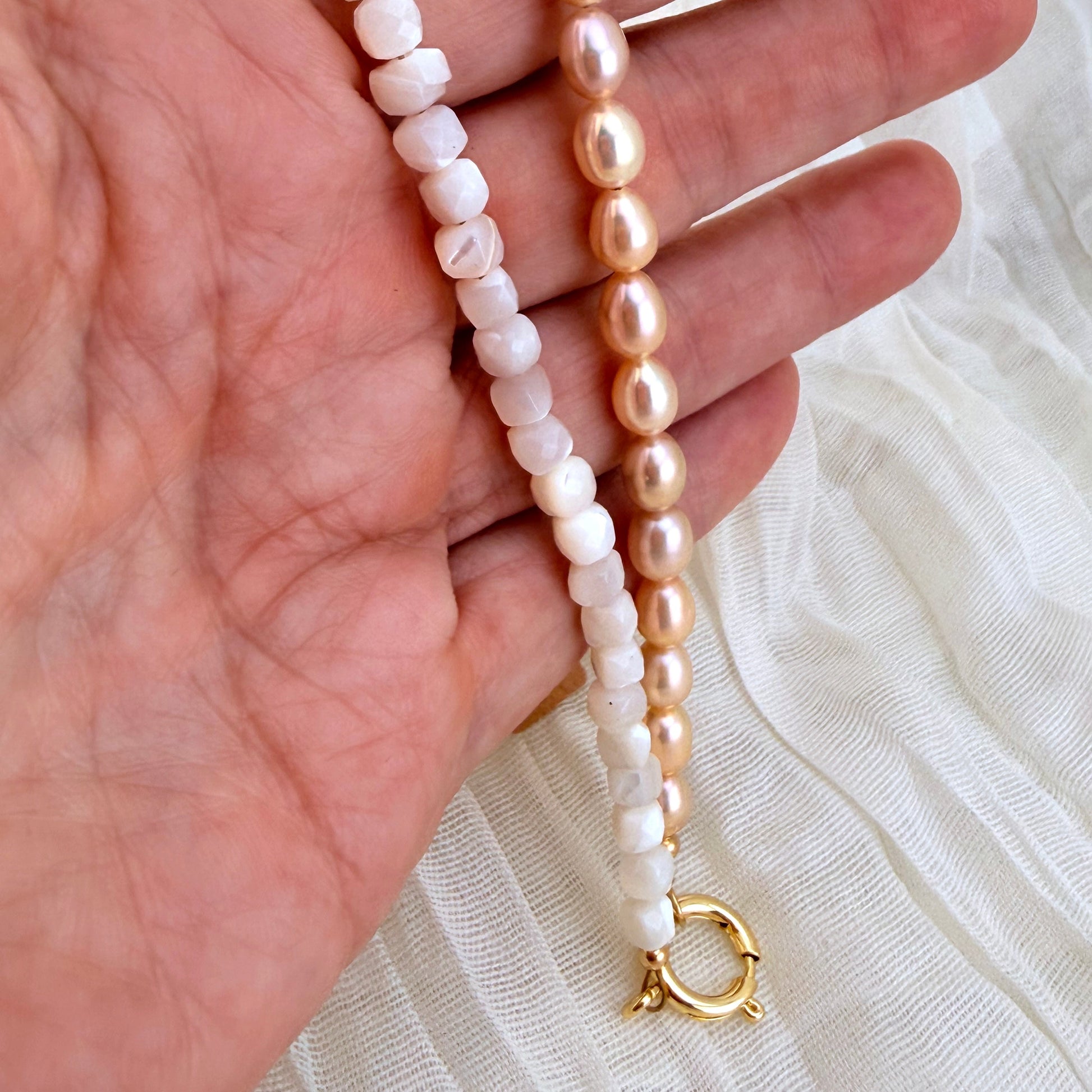 Two beaded strands draped over a hand: one strand features  white beads with gold settings, and the other features smooth, oval light pink pearls with gold settings. Both strands terminate in a gold clasp.