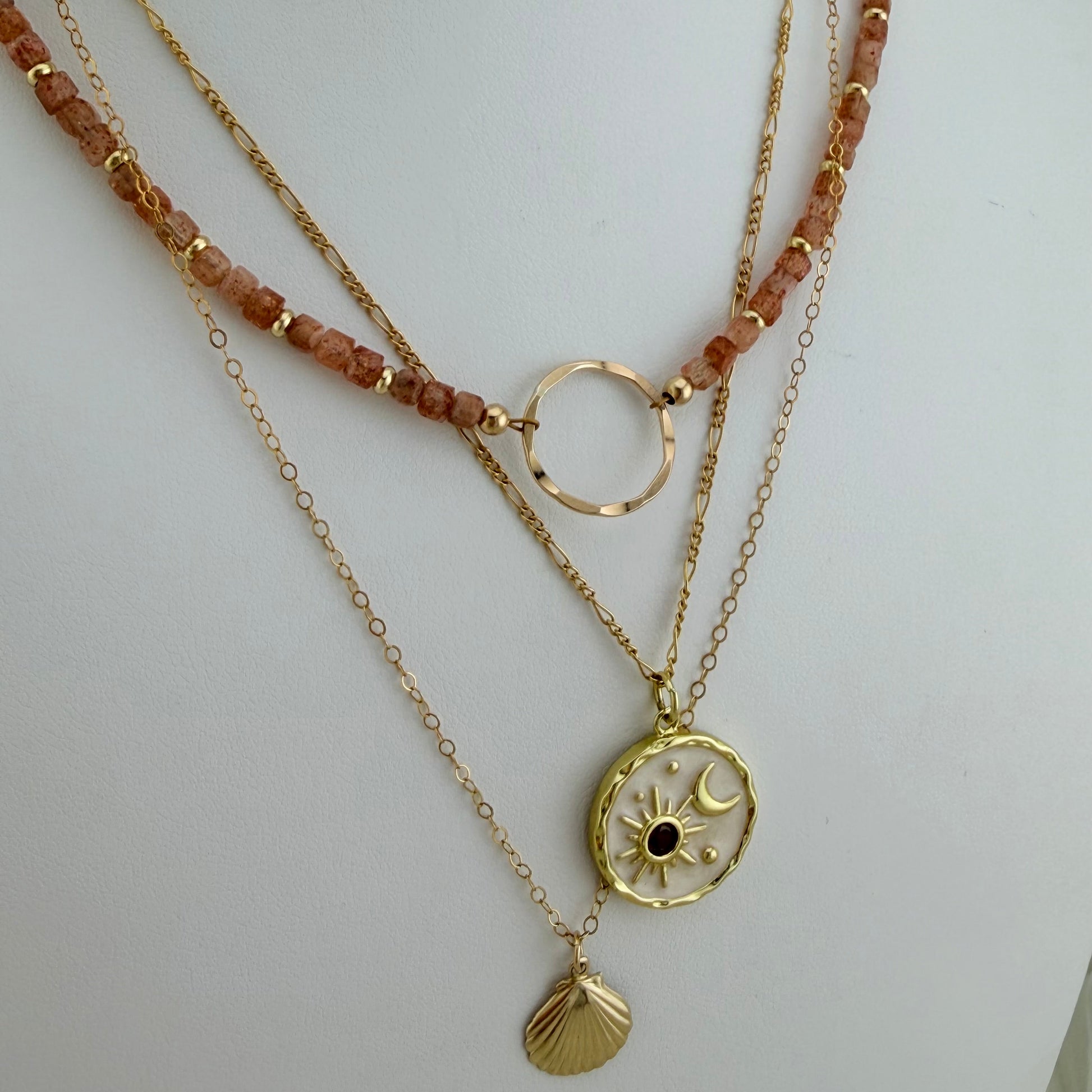 A layered gold necklace set displayed on a white mannequin bust. The necklace features a beaded strand with peach-colored stones, a gold ring connector, and three dangling chains: one with a gold sun and moon pendant, one with a small gold shell pendant, and one with a thin chain.